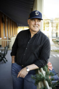 Promotional photo of Ace Atkins leaning on a balcony rail wearing a baseball cap